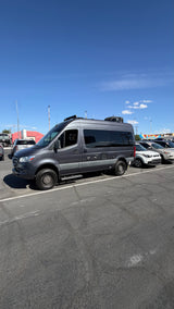An adventure-ready grey Mercedes Sprinter rv rental, equipped with rugged tires, is parked outdoors under a blue sky.
