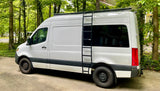 White Mercedes Sprinter RV rental van with roof rack & ladder, parked in a lush, green setting.
