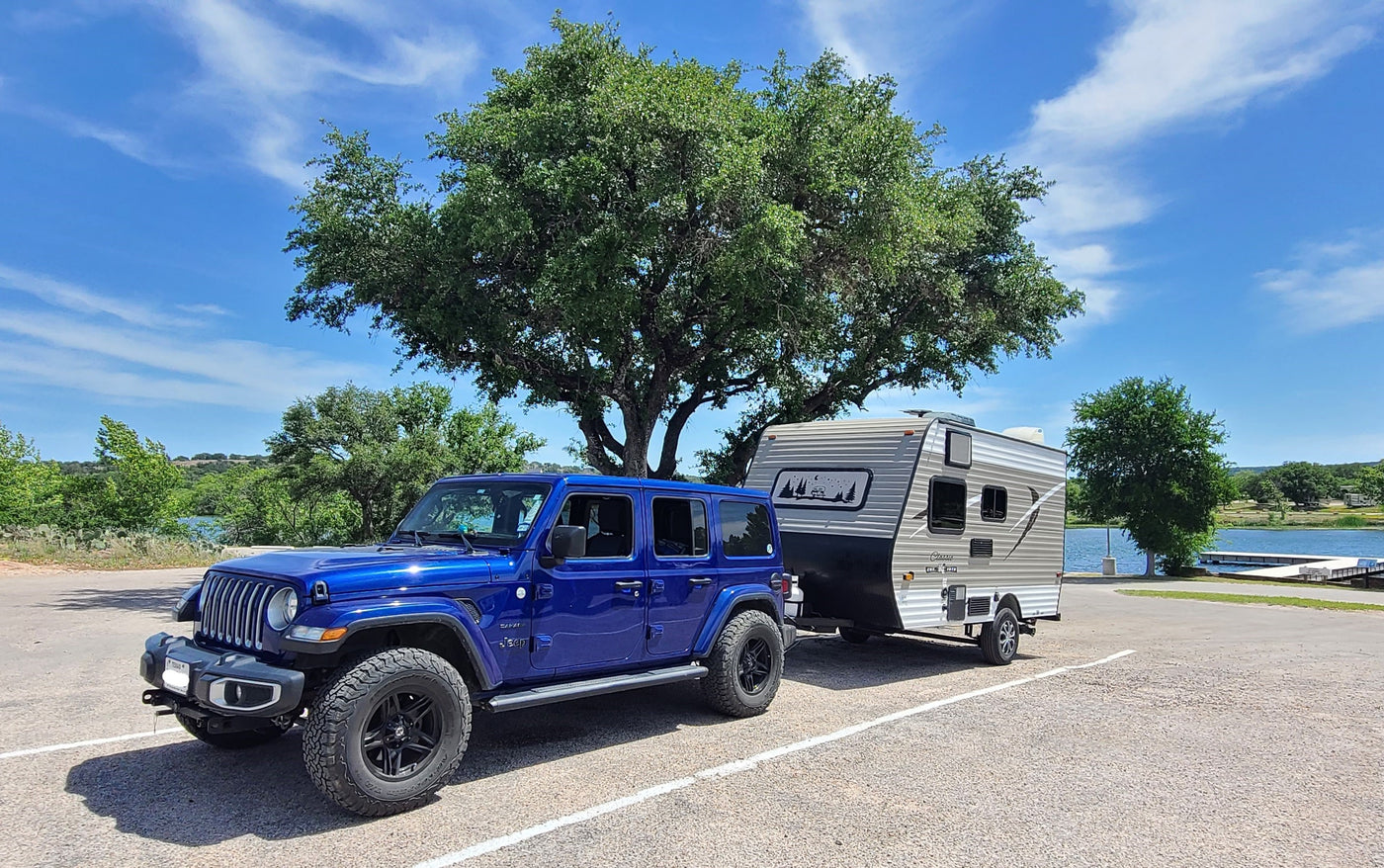 Blue Jeep Wrangler towing a compact RV rental by a lake, ideal for camping adventures in nature.
