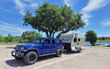Blue Jeep Wrangler towing a compact RV rental by a lake, ideal for camping adventures in nature.