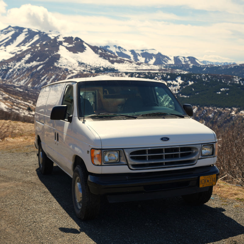 A white cargo van is parked on a gravel mountain road with snow-capped peaks in the background. Ideal for an Alaska RV rental adventure. — photo 2