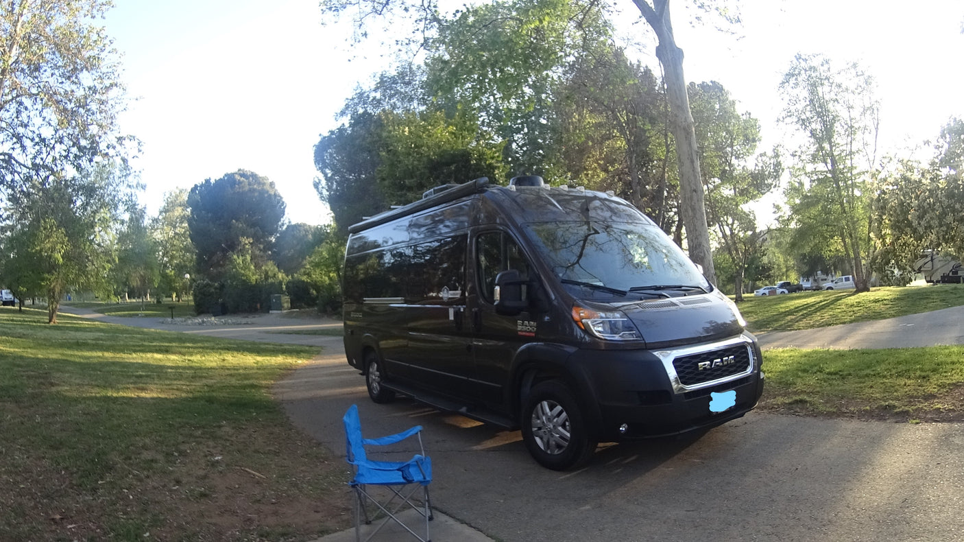 Dark grey Ram ProMaster RV rental van parked on a path in a sunny park with trees and a blue chair.