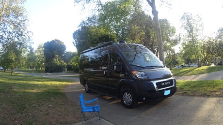 Dark grey Ram ProMaster RV rental van parked on a path in a sunny park with trees and a blue chair.