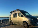 A grey Storyteller Overland Sprinter van RV rental is parked on a paved lot beside a sandy beach and ocean.