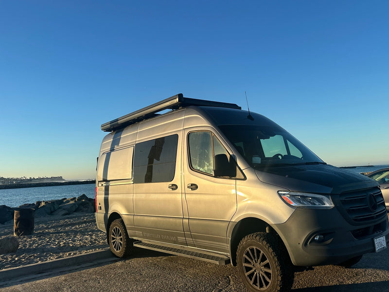 A grey Storyteller Overland Sprinter van RV rental is parked on a paved lot beside a sandy beach and ocean. — photo 1