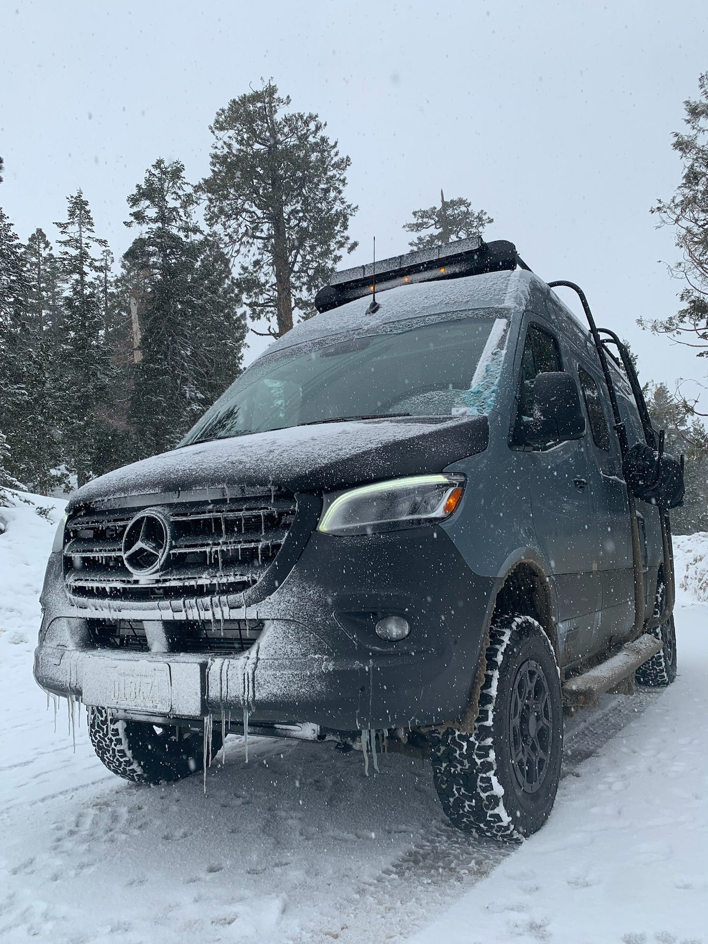 Snowy Mercedes-Benz Sprinter RV rental in a winter forest, covered in snow & icicles, ready for off-road adventure.