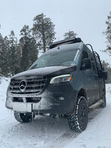 Snowy Mercedes-Benz Sprinter RV rental in a winter forest, covered in snow & icicles, ready for off-road adventure.