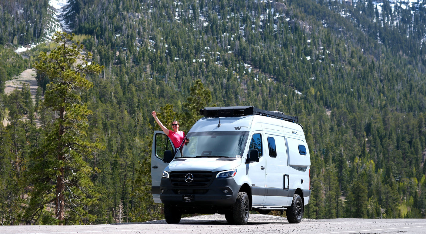 Explore mountains with an rv rental! A woman waves from a gray Winnebago Revel Sprinter van amidst pine forests.