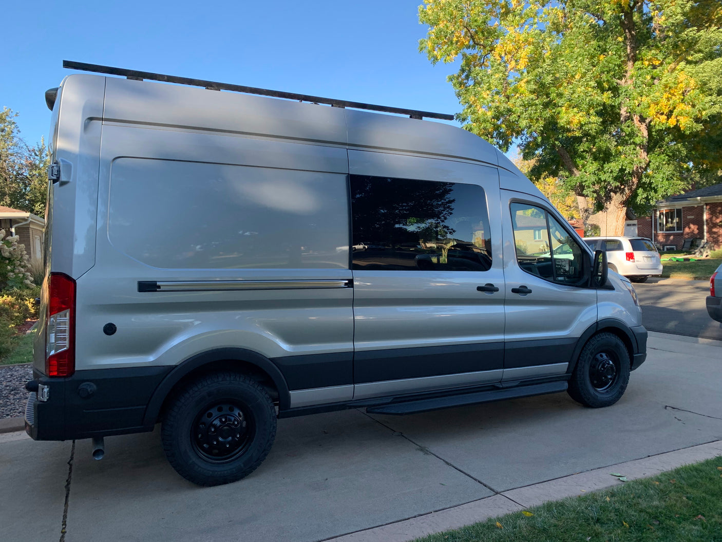 Adventure-ready silver Ford Transit camper van for RV rental, parked on a driveway, with roof rack and rugged tires.