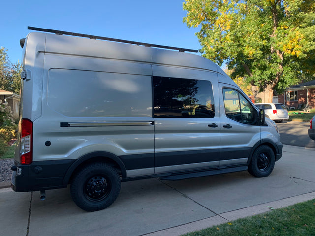 Adventure-ready silver Ford Transit camper van for RV rental, parked on a driveway, with roof rack and rugged tires.