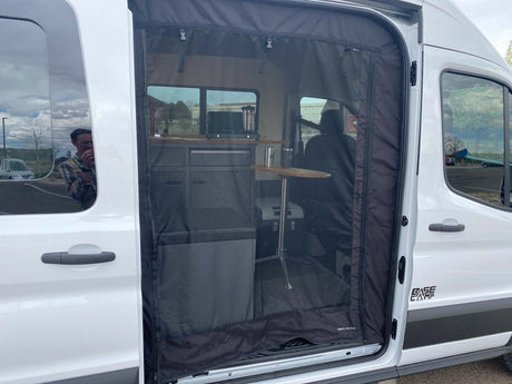 RV rental camper van interior. Side door open with bug screen, revealing a compact kitchen, table, and seating.
