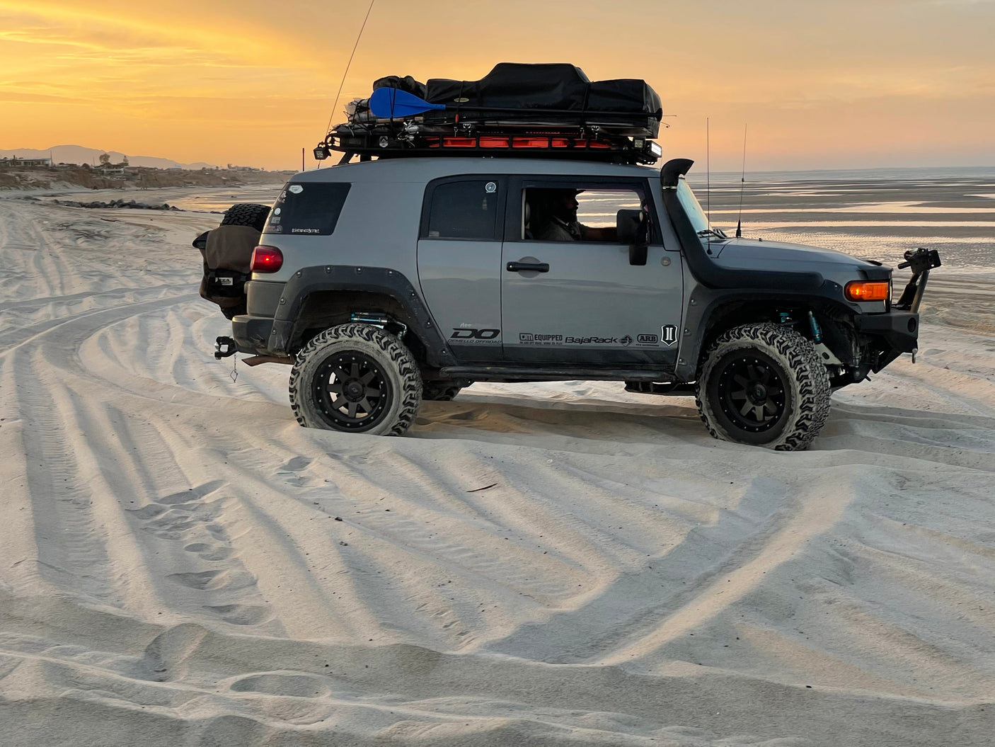 Gray modified FJ Cruiser with rooftop tent on sandy beach at sunset. Ideal for adventure travel; consider this like an RV rental!