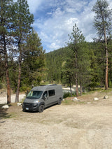Gray camper van RV rental parked by a river in a mountainous forest setting under a cloudy blue sky.