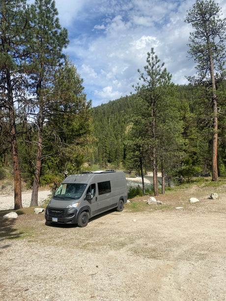 Gray camper van RV rental parked by a river in a mountainous forest setting under a cloudy blue sky.