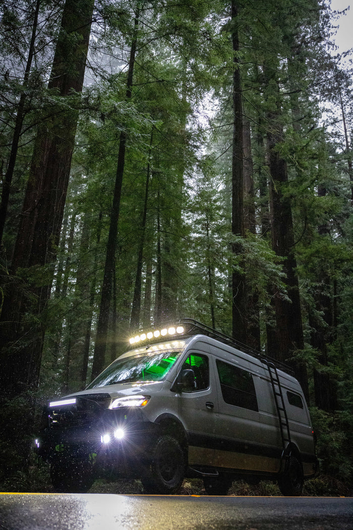 Off-road RV rental van lighting up a misty redwood forest road, ready for adventure among towering trees.