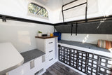 Bright RV rental interior showing a compact dinette, kitchen counter with drawers, and multi-functional seating that converts to a bed. Modern design.