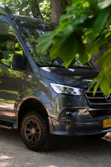 Dark gray Mercedes Sprinter camper van, an RV rental, with off-road tires, parked outdoors beneath leafy trees.
