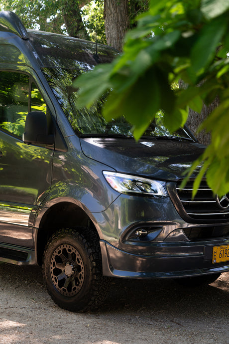 Dark gray Mercedes Sprinter camper van, an RV rental, with off-road tires, parked outdoors beneath leafy trees.