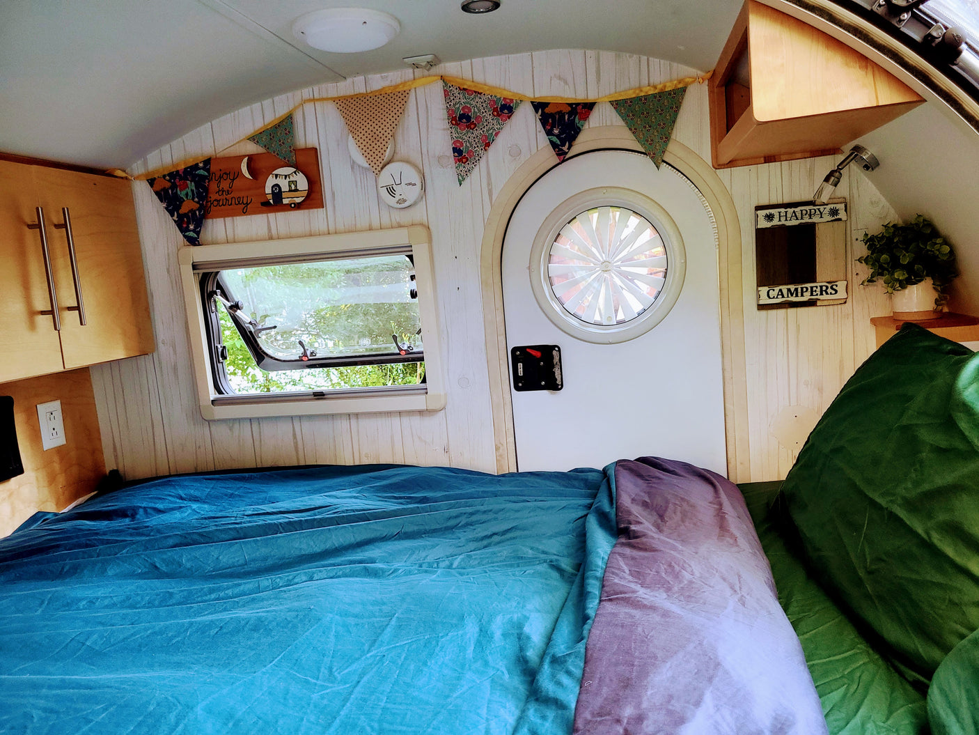 Inside a cozy RV rental: a bed with blue and purple covers, an open window to nature, and charming camper decor. Ready for travel!