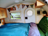 Inside a cozy RV rental: a bed with blue and purple covers, an open window to nature, and charming camper decor. Ready for travel!