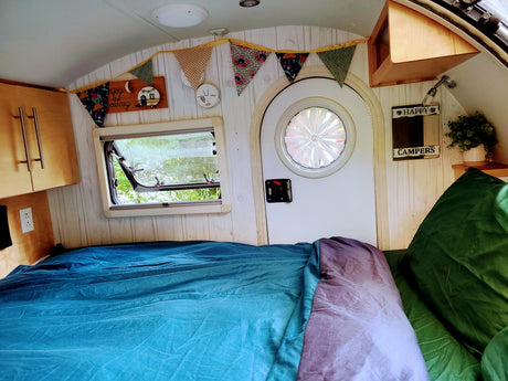 Inside a cozy RV rental: a bed with blue and purple covers, an open window to nature, and charming camper decor. Ready for travel!