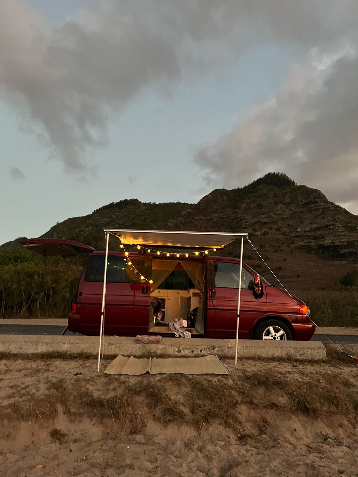 Red camper van RV rental with illuminated awning set for cozy evening camping by sandy beach