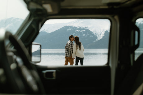 A couple kisses by a mountain lake, seen through the window of an RV rental. Snowy peaks in background.