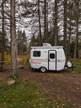 White Scamp travel trailer, a compact RV rental, awaits adventure in a peaceful autumn forest campsite.