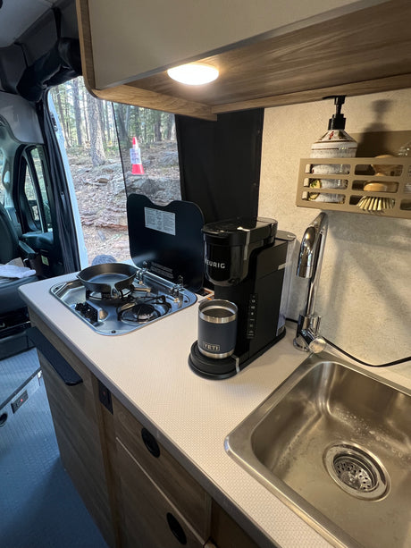 RV rental kitchenette with a stove, Keurig coffee maker, sink, and a scenic forest view through the window.