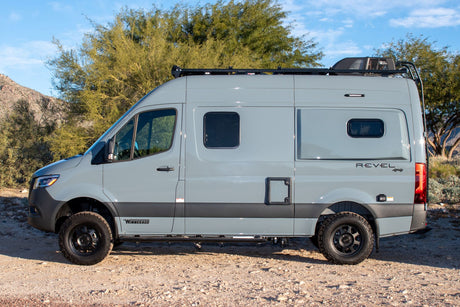 Side view of a gray Winnebago Revel 4x4 camper van parked on a dirt road. Ideal for an RV rental adventure.