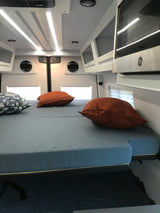 Modern RV rental interior: A bright, comfortable bed with pillows, overhead LED lighting, cabinets, and a microwave.