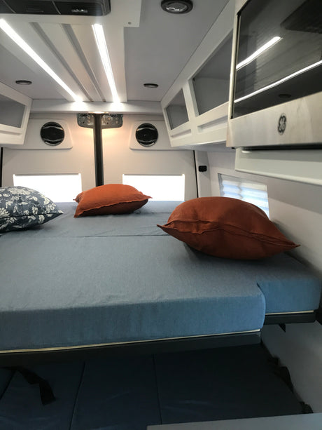 Modern RV rental interior: A bright, comfortable bed with pillows, overhead LED lighting, cabinets, and a microwave.