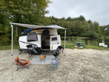 Cozy RV rental setup by a serene lake. A compact trailer with an awning, chairs, and table offers outdoor comfort.