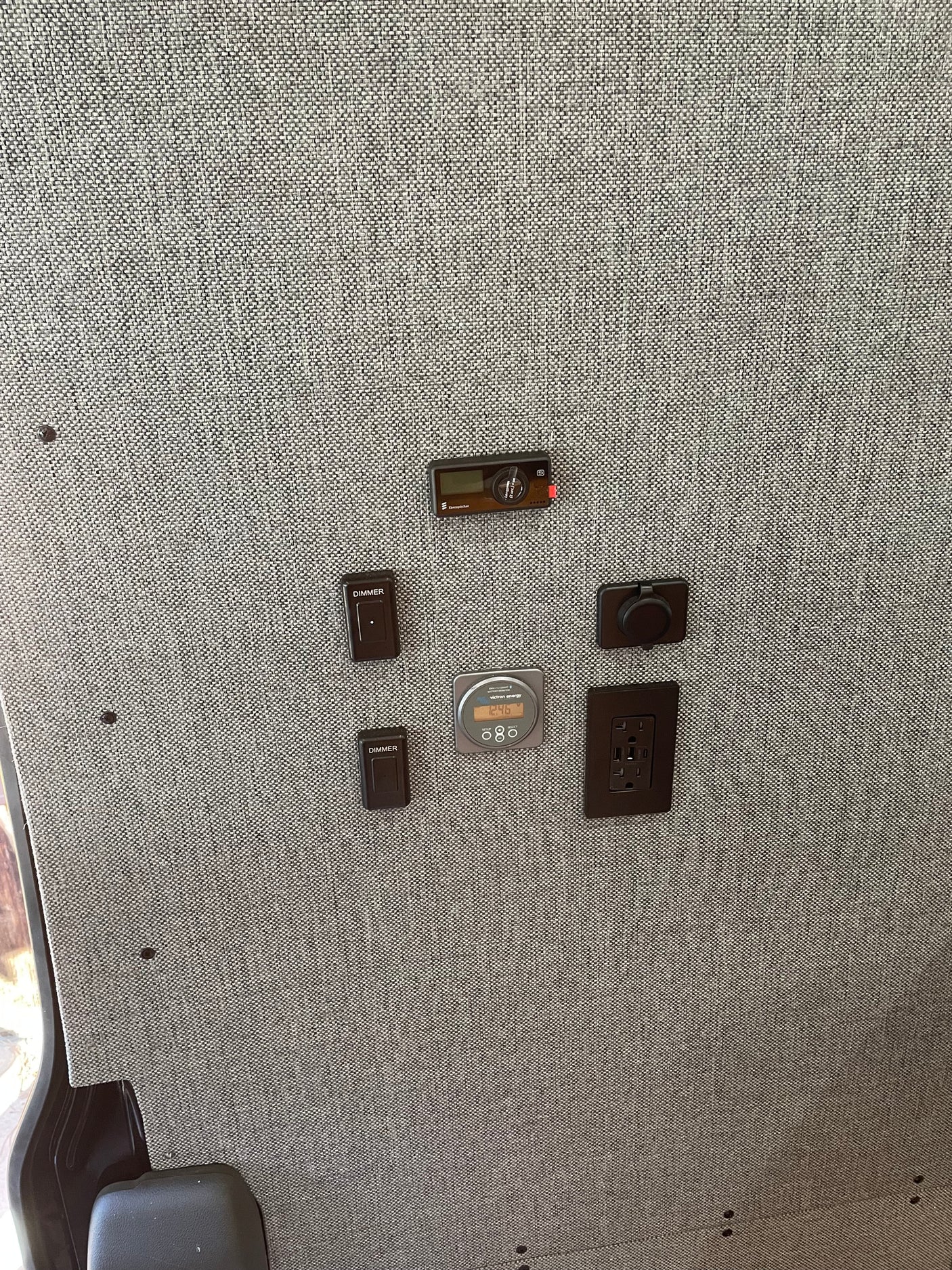 RV rental interior featuring various controls: heater, dimmers, battery monitor, and outlets on a textured gray fabric wall.