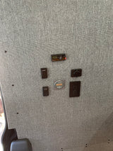 RV rental interior featuring various controls: heater, dimmers, battery monitor, and outlets on a textured gray fabric wall.