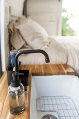 Inside an RV rental: a black faucet, clear soap dispenser, and white sink on a wooden counter, with a cozy bed in the background.