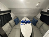 Inside an RV rental: a cozy U-shaped dinette with grey cushions, a white table set with blue dishes, and black window blinds.