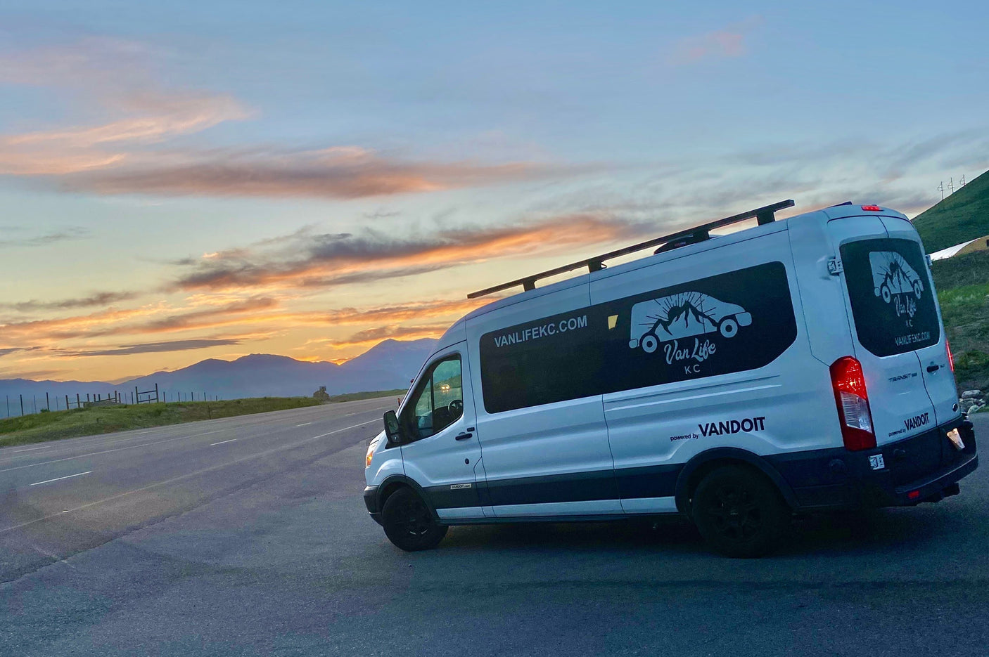 A white Van Life KC RV rental, a Ford Transit camper van, parked overlooking mountains at sunset. Ready for adventure.