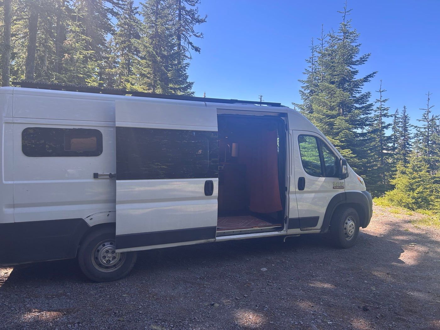White RV rental camper van with an open door, parked on a sunny gravel path among tall evergreen trees under a clear blue sky. — photo 2