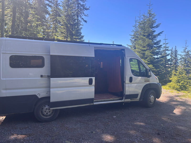 White RV rental camper van with an open door, parked on a sunny gravel path among tall evergreen trees under a clear blue sky. — photo 2