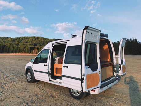 White camper van (RV rental) with doors open on a sandy shore. Inside, a cozy, custom-built bed. Scenic forest view.