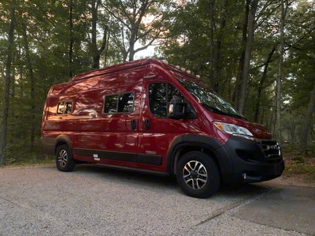 This red RV rental camper van is parked on gravel amidst lush trees, ready for a serene nature road trip adventure.