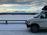 A white van (possible rv rental) with a roof box on a snowy road, facing a vast snowfield and distant Alaskan mountains under cloudy skies.