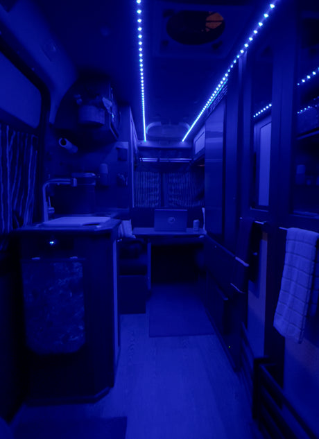 Experience mobile living in this blue-lit RV rental interior, featuring a compact kitchenette and a laptop workspace.