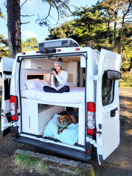 Cozy RV rental adventure: Woman sips coffee on bunk bed while dog rests below in a scenic camper van.