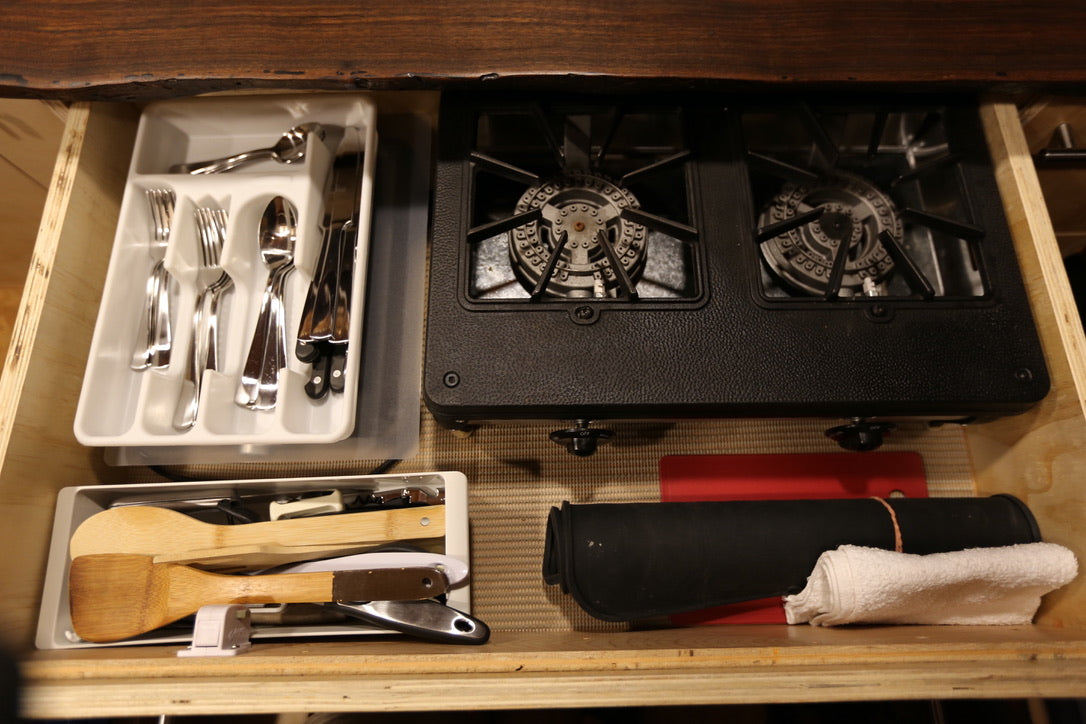 RV rental kitchen drawer features a compact stove, organized cutlery, and wooden cooking utensils.