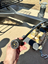 A hand holds a 7-pin trailer electrical connector, vital for RV rental towing. Trailer frame and safety chains are in the background.