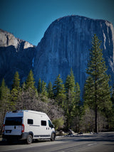 White RV rental parked on a road, dwarfed by a massive granite mountain and surrounded by tall pine trees.