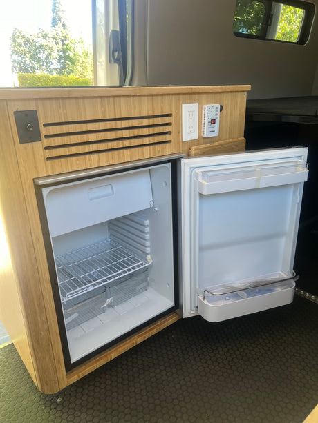 An RV rental interior with a built-in, open refrigerator/freezer in bamboo cabinetry, plus power outlets & a monitor.