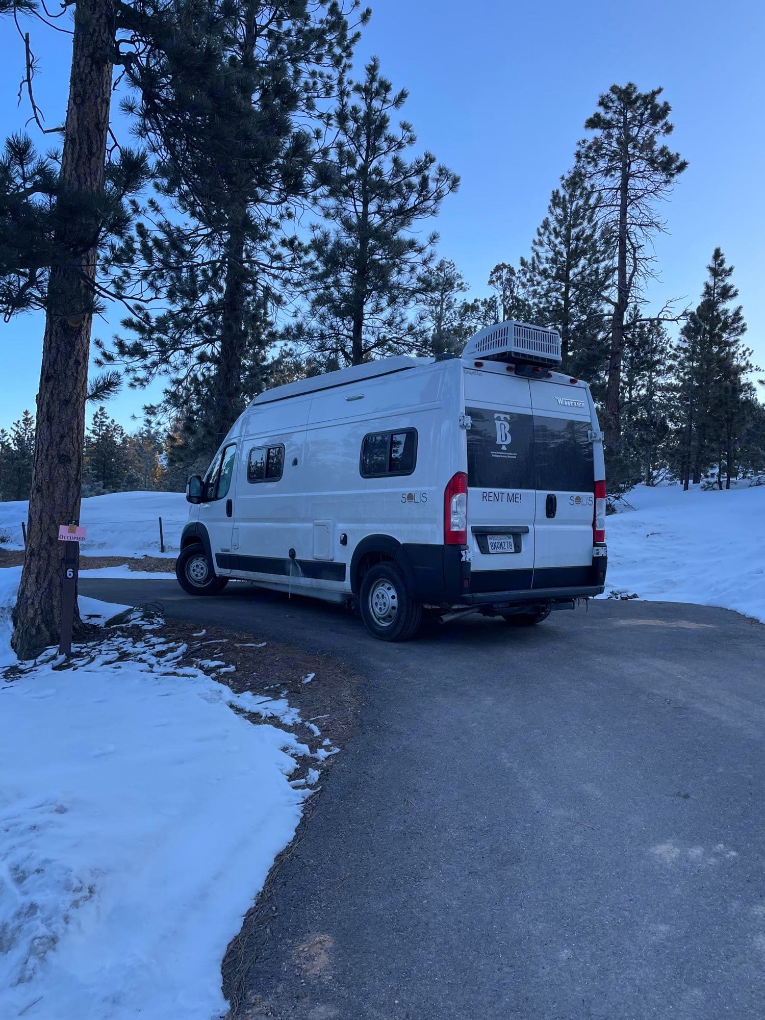 White Winnebago Solis RV rental parked in a snowy forest campsite. Ideal for winter adventures.